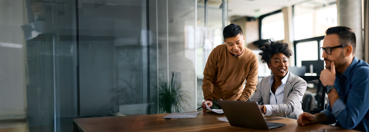 Three diverse professionals collaborating in a modern office, discussing work around a laptop.
